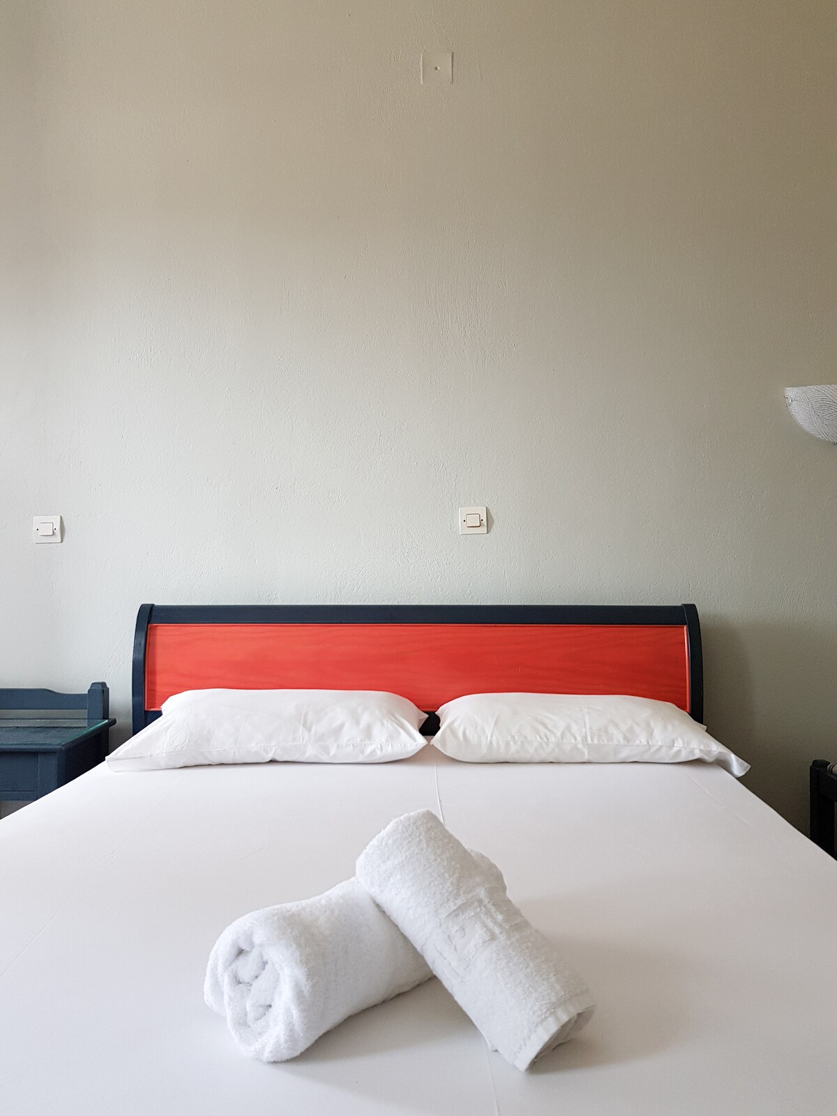 A comfortable sleeping area is presented with a neatly made bed, featuring a red headboard and crisp white linens. Two rolled towels are placed at the foot of the bed. Light-colored walls enhance the bright and airy feel of the space.