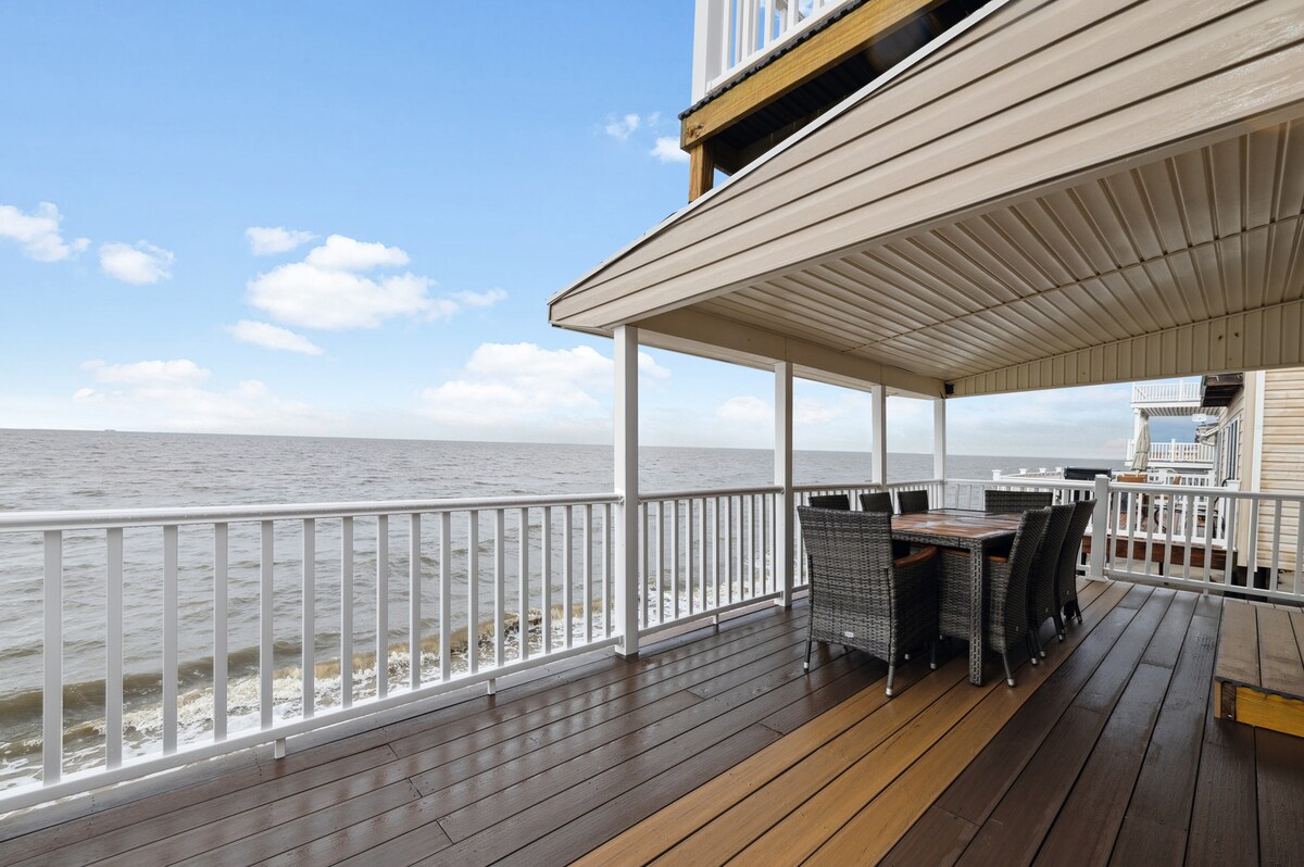 A spacious deck is presented, featuring a dining table with six chairs positioned under a covered area. The expansive bay view stretches out, with gentle waves lapping at the shore, creating a serene backdrop for outdoor meals and relaxation.