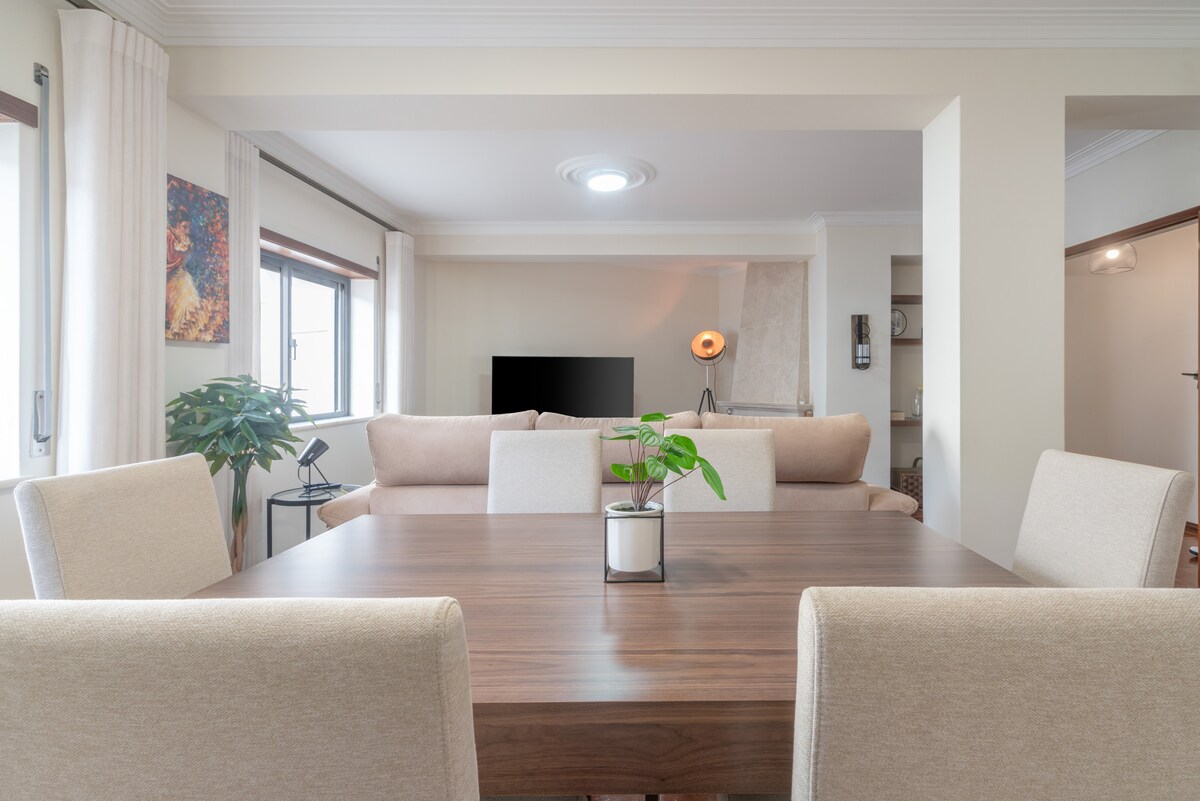 A modern dining area features a large wooden table surrounded by beige upholstered chairs. A small potted plant sits at the center. Natural light enters through windows, illuminating the open space that connects to a cozy living area with a sofa and a television.