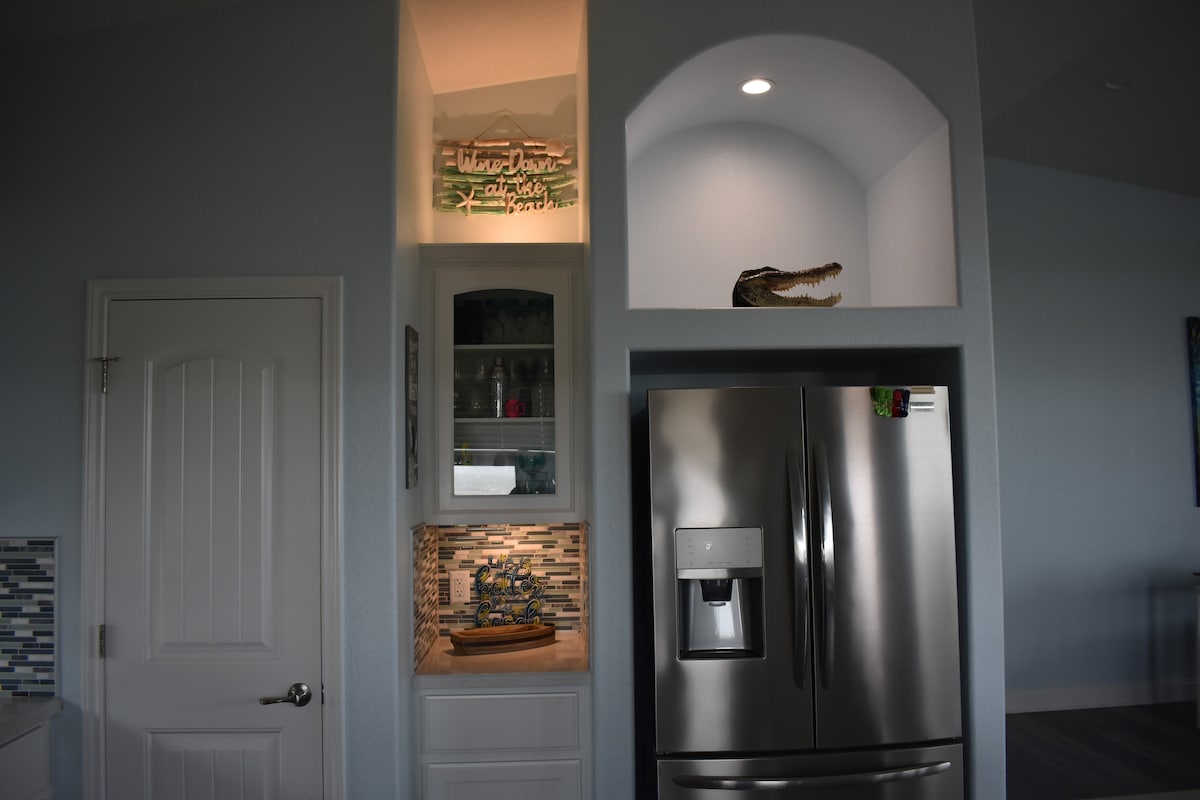 A modern kitchen features a stainless-steel refrigerator alongside white cabinetry. A glass-front cabinet showcases decorative items, while a small alcove holds a crocodile sculpture. The backsplash is adorned with tiles in shades of blue and gray, adding subtle texture to the space.