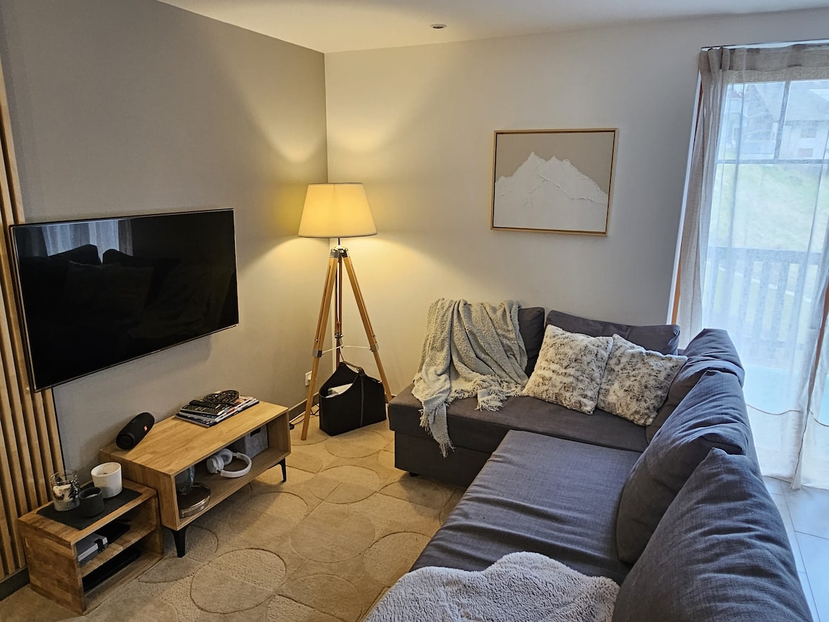 A cozy living area is visible, featuring a sectional sofa adorned with soft throw blankets and decorative pillows. A wooden coffee table holds a few items, while a floor lamp and wall-mounted television enhance the space. Natural light enters through sheer curtains, creating a bright atmosphere.