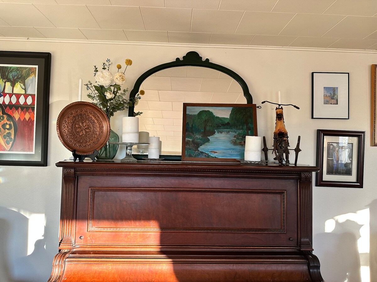 An antique wooden piano serves as a focal point, accompanied by a collection of framed artwork and photographs. A large mirror with a dark green frame hangs above the piano, while decorative elements include candles and a vase with flowers, enhancing the overall aesthetic.