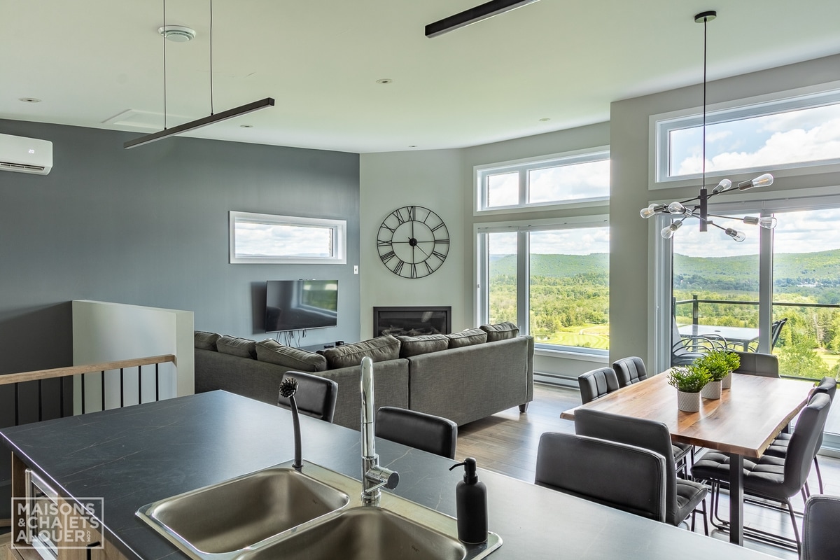 An open-concept living space showcases a modern kitchen with a sleek sink, leading to a dining area featuring a wooden table surrounded by black chairs. Large windows provide views of the green landscape outside, while a cozy seating area is arranged for relaxation.