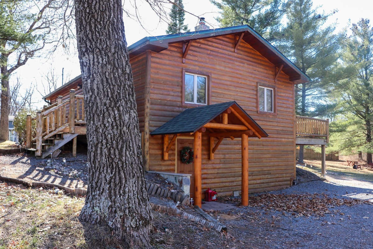A rustic cabin features a wooden exterior with a welcoming porch entry. The structure is surrounded by trees, and a spacious deck is visible above, complemented by natural gravel pathways. Sunlight creates soft shadows on the ground, enhancing the natural setting.