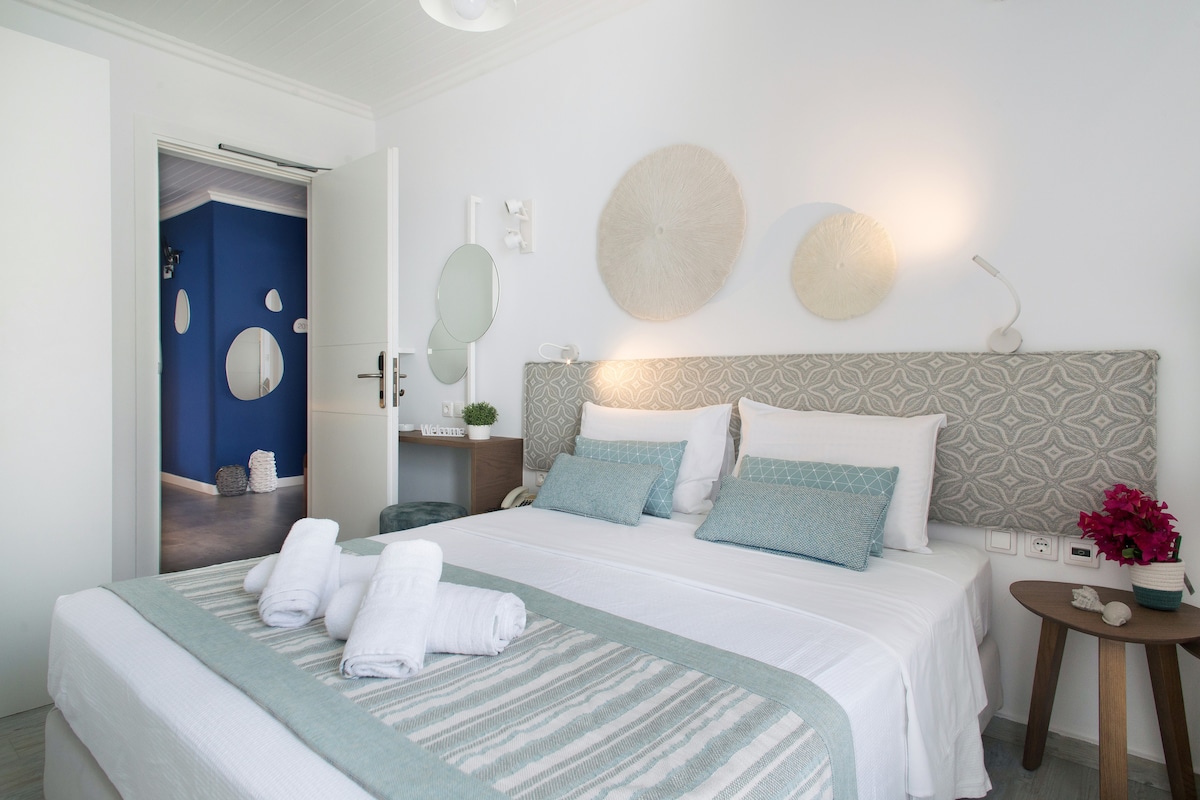 A modern room features a spacious bed with a geometric headboard, adorned with decorative pillows in soft blue tones. Natural light illuminates the area, highlighting the textured wall art. A small bedside table and wall-mounted lighting provide functionality. A doorway visible leads to another room.