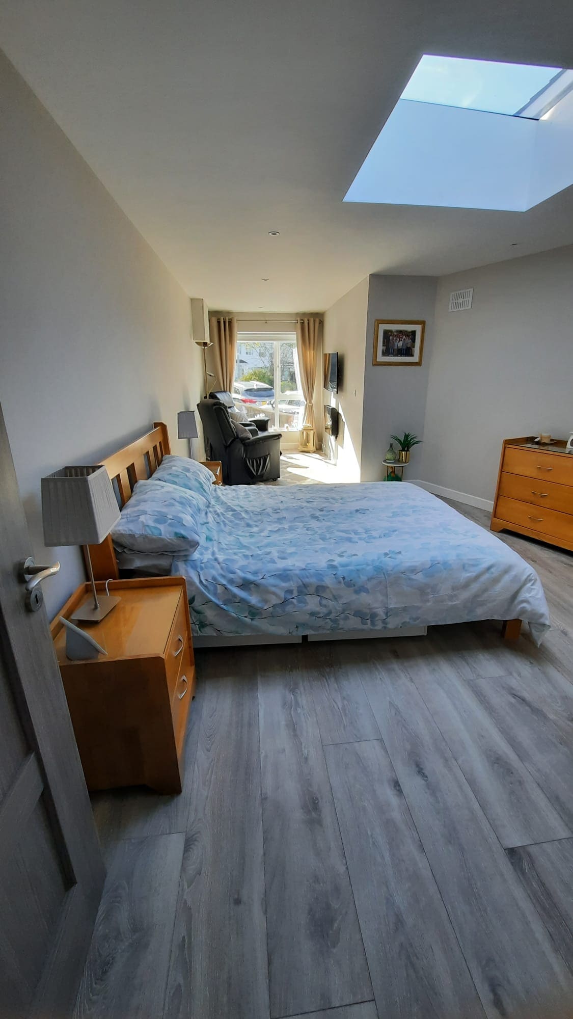 A comfortable room features a spacious bed covered in a light-colored duvet. A wooden nightstand and dresser are present, while natural light fills the space through a skylight and nearby window, creating an inviting atmosphere.