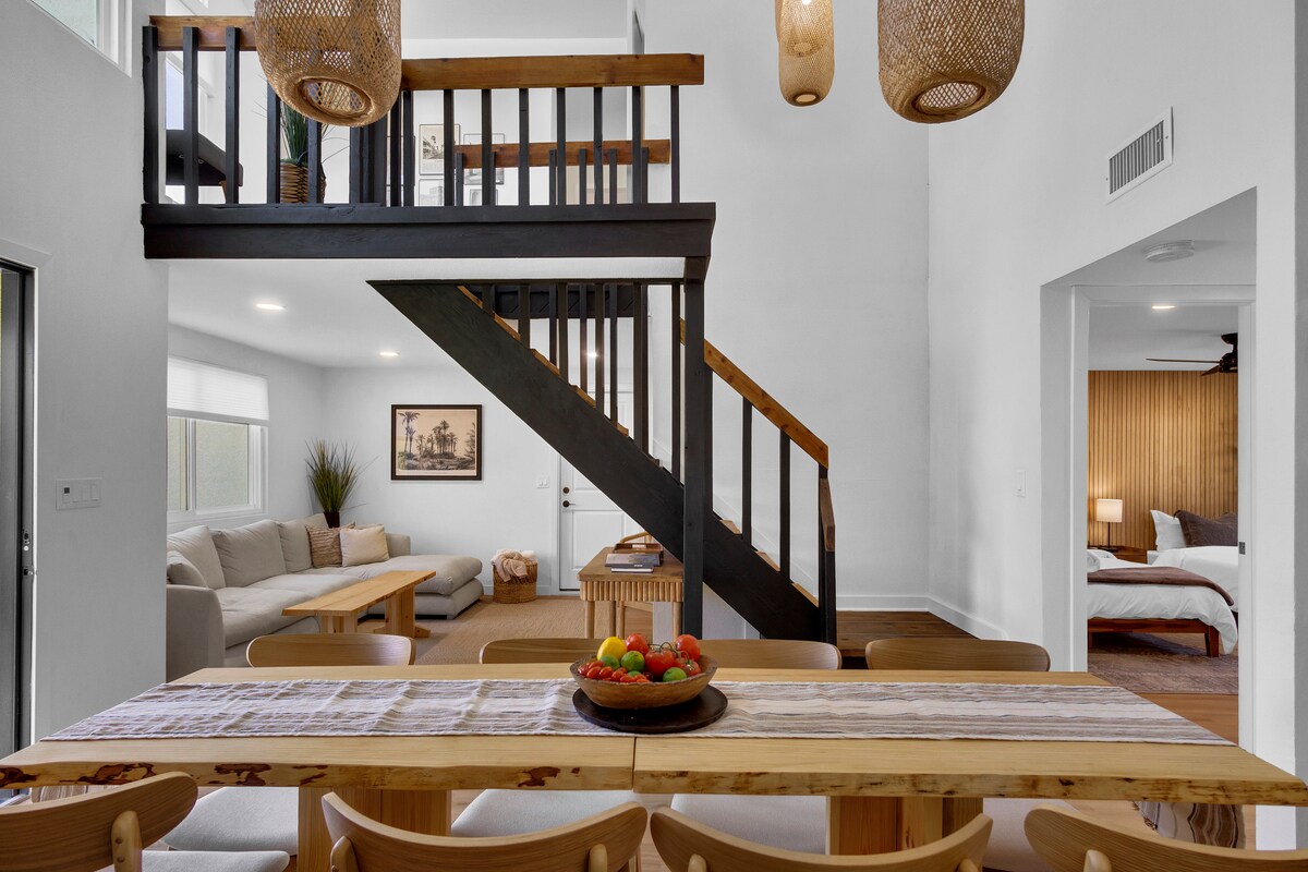 A spacious dining area features a long wooden table surrounded by multiple chairs, inviting family meals. An open staircase leads to a lofted space, while a cozy living area with a sectional sofa can be seen in the background, enhancing the home's welcoming atmosphere.