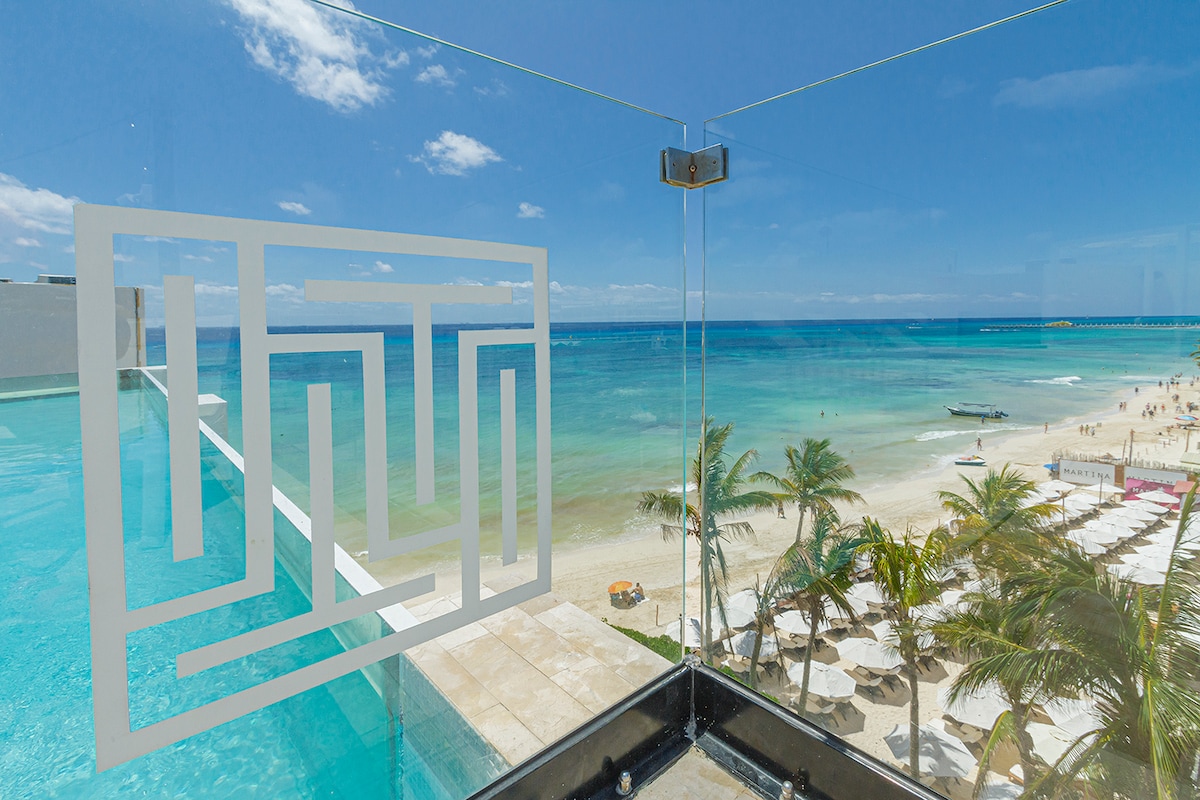 A panoramic view of the Caribbean Sea is captured from the balcony, showcasing clear turquoise waters and sandy beaches. Palm trees are visible in the foreground, while the rooftop pool reflects the vibrant blue sky above.