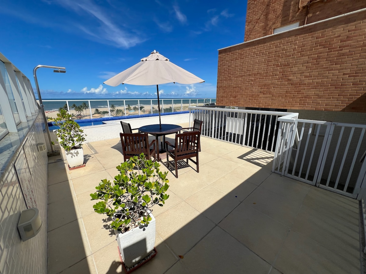 A spacious rooftop area features a round table surrounded by four wooden chairs, shaded by a large umbrella. Potted plants add greenery to the space, which overlooks a swimming pool and offers views of the beach and sky.