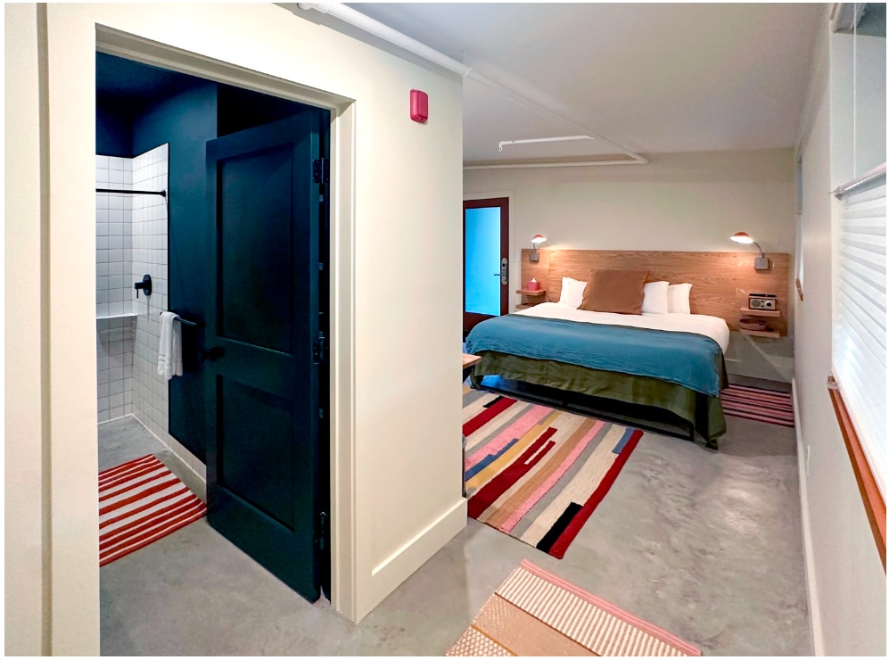 A spacious room is visible, featuring a king bed with a custom headboard. A stylish writing desk is located beside the bed, along with a cozy sitting chair. The entryway leads to a modern bathroom with a tiled walk-in shower, enhancing the room's aesthetic.