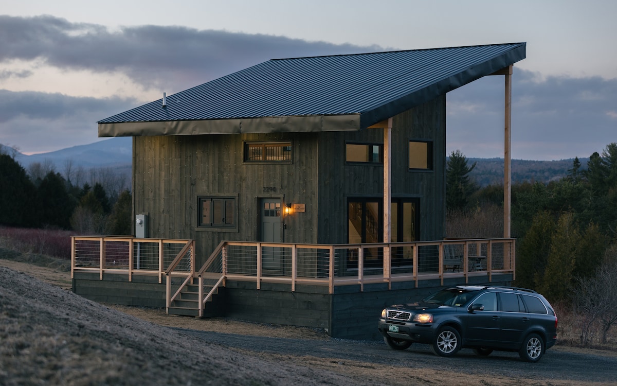 The exterior of a modern cabin features a sloped metal roof and large windows, allowing natural light to enter. A wooden deck wraps around the front, supported by sturdy posts. A vehicle is parked nearby, with rolling hills and trees in the background.