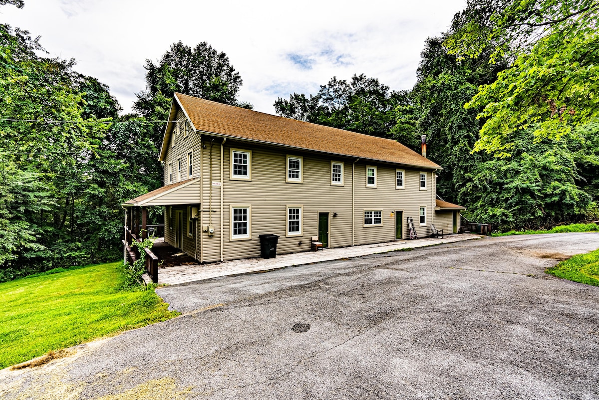 A large, two-story retreat center is set amidst lush greenery, featuring a welcoming front porch and multiple windows. A paved driveway leads to the building, with ample parking space visible. The natural surroundings enhance the property’s tranquility.