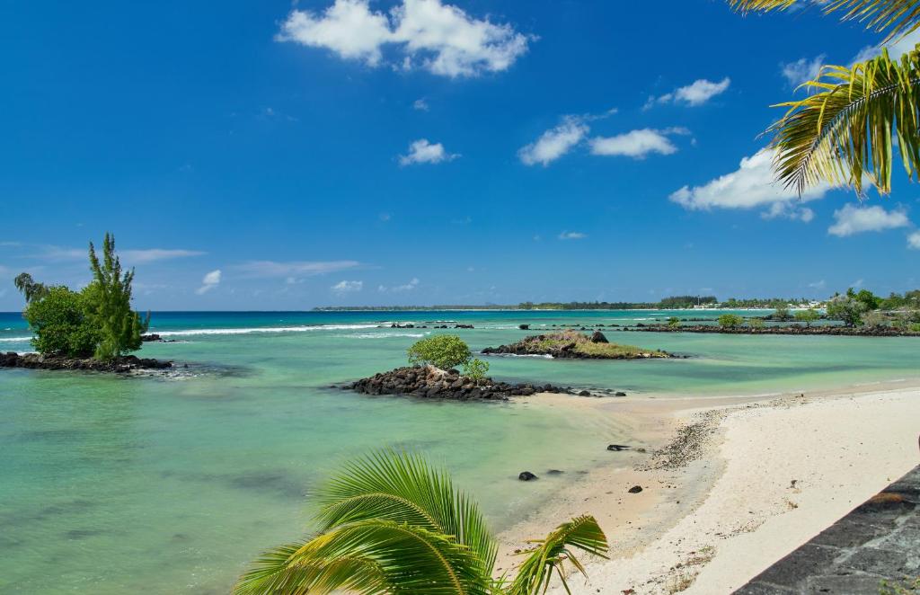 The image captures a scenic view of a tranquil shoreline bordered by soft, white sand and clear turquoise waters. Lush greenery and small rocky outcrops are visible amidst the gentle waves, while a bright blue sky dotted with clouds enhances the serene coastal environment.
