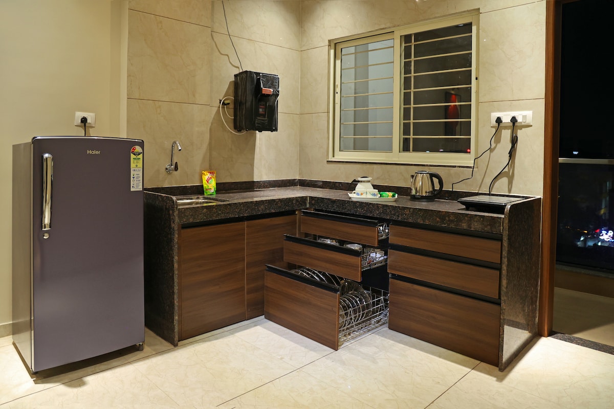 A modern kitchen features a sleek refrigerator, a granite countertop, and a minimalist sink. Dark wooden cabinets are present, with several drawers partially opened to reveal storage options. Natural light enters through a window, illuminating the space.