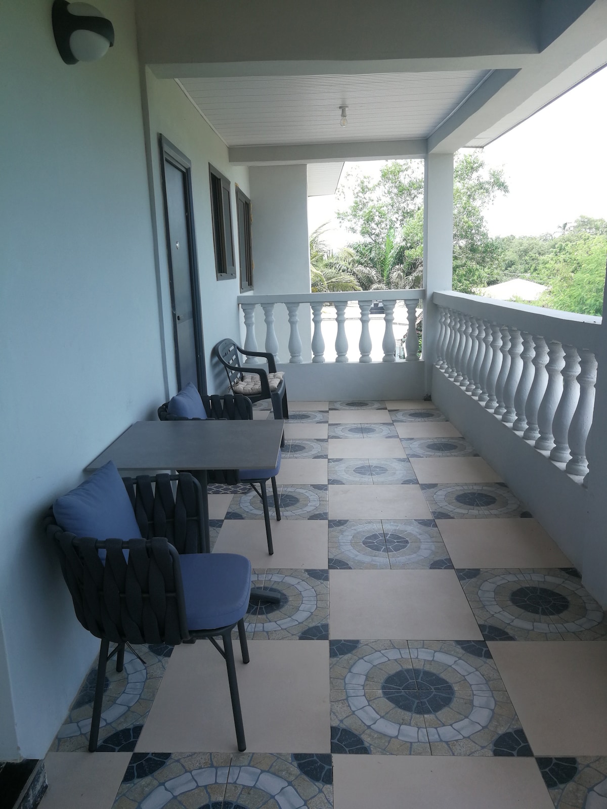 A balcony area is visible, featuring a patterned tile floor and a white railing. Two black chairs and a small table are positioned against the wall, offering a space for relaxation. Natural light floods the area, enhancing the view of the surrounding greenery.