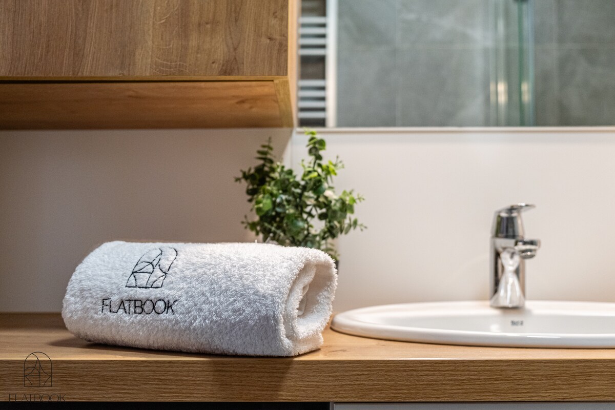 A white towel with an embroidered logo rests neatly on a wooden countertop. A small green plant adds a touch of nature, while a sleek faucet is positioned near a circular sink set against a backdrop of gray tiles.