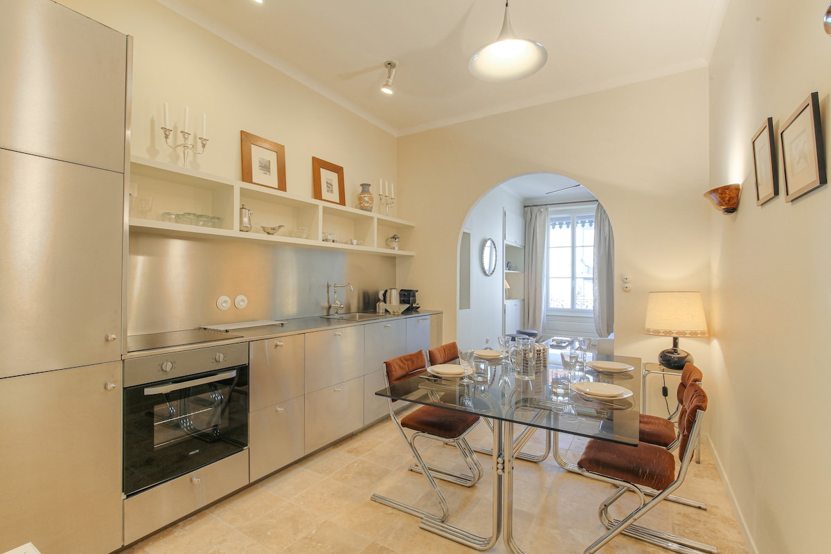 A sleek kitchen area features stainless steel cabinetry and appliances, with light-colored walls enhancing the brightness. A glass dining table is surrounded by contemporary chairs. Natural light streams through an archway leading into a comfortable living space, indicated by a glimpse of soft furnishings.
