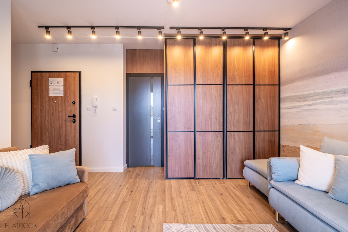 A modern living space is presented, featuring a light wooden floor and a combination of soft furnishings in neutral tones. A versatile storage unit with wood paneling serves as a backdrop, while warm lighting creates a comfortable ambiance. The entryway door is visible on one side.