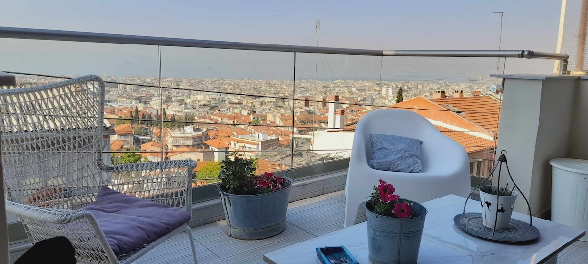 A balcony is shown featuring a seating area with a white chair and a cushioned chair, providing a space for relaxation. Potted plants with vibrant flowers add color, while a small table holds decorative items. The expansive view captures the rooftops of the city below.