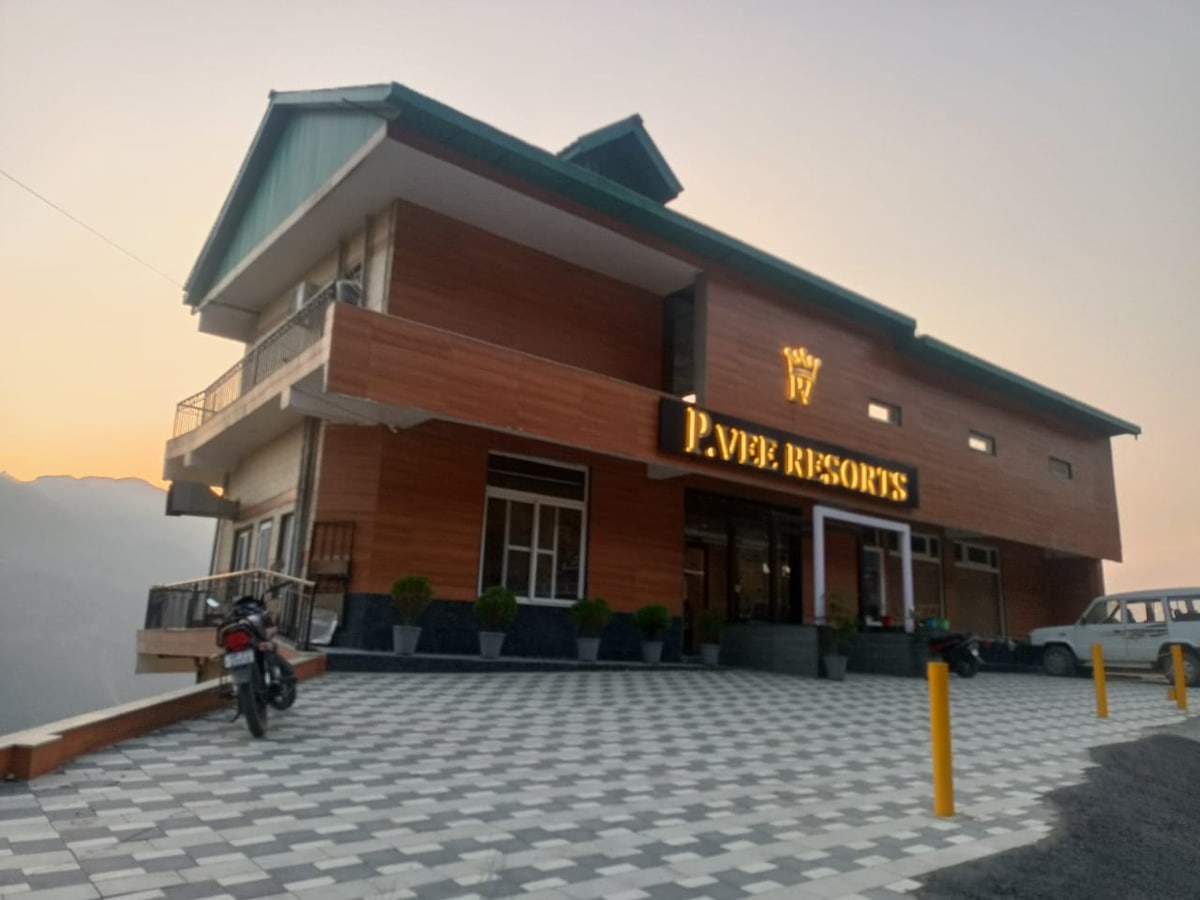 The exterior of P.Vee Resorts is shown, featuring a modern wooden facade and expansive glass windows. A paved parking area is visible, with plants lining the entrance. The building is set against a backdrop of gentle hills and a soft evening sky.