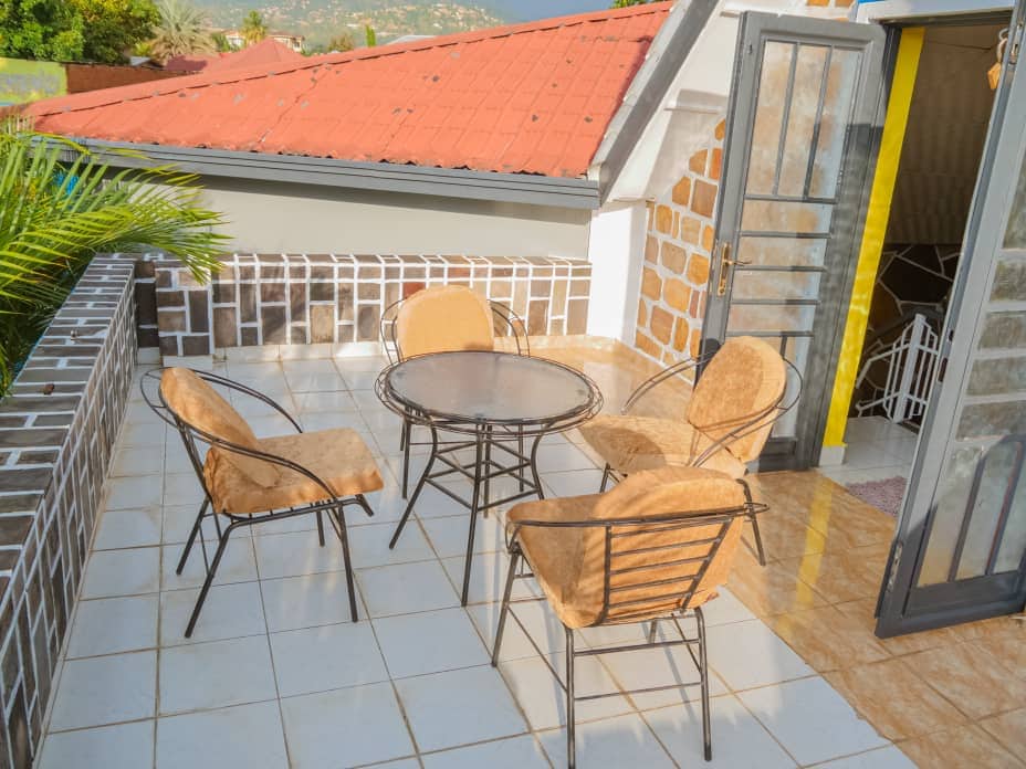 An outdoor balcony area is equipped with a round table and four chairs, positioned on tiled flooring. The walls are adorned with a mix of stone and painted surfaces, complemented by a view of surrounding rooftops and greenery.
