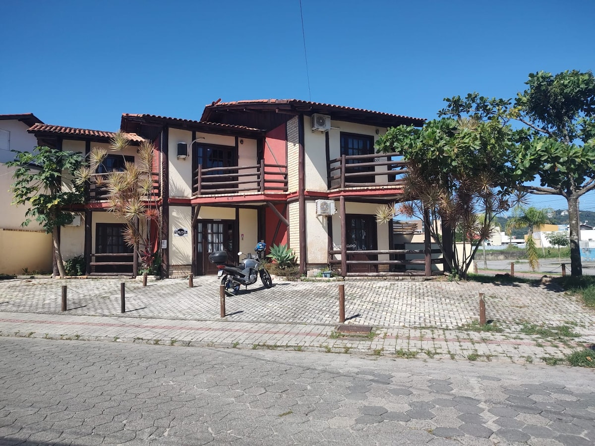 A two-story building is displayed, featuring a combination of red and beige walls. Balconies with wooden railings provide an outdoor view. The front area includes a paved seating space with palm trees and a motorcycle parked near the entrance.