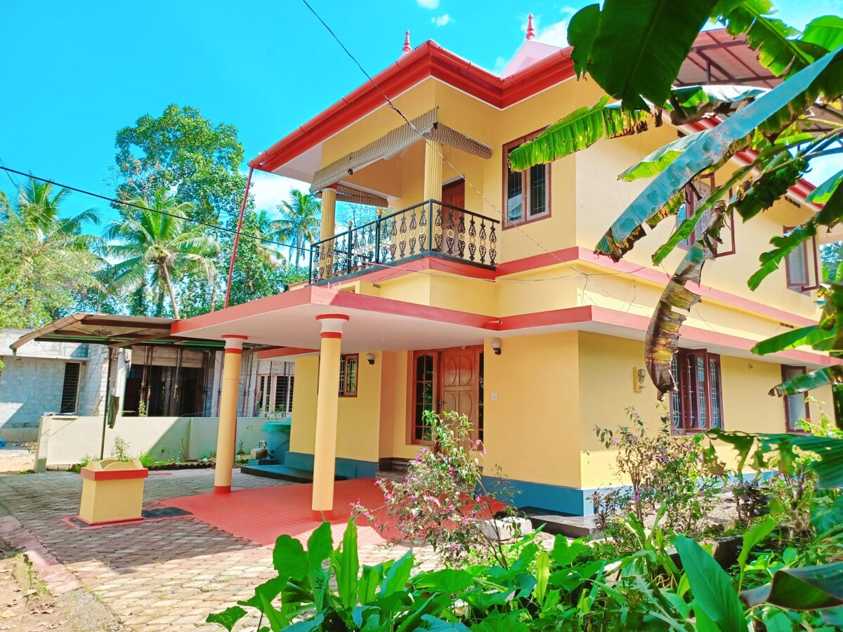 A charming rustic villa features a bright yellow exterior, highlighted by a red roof and balcony. Lush green plants frame the entrance, while palm trees provide a tropical backdrop. The pathway is lined with colorful foliage, inviting a sense of tranquility amidst nature.