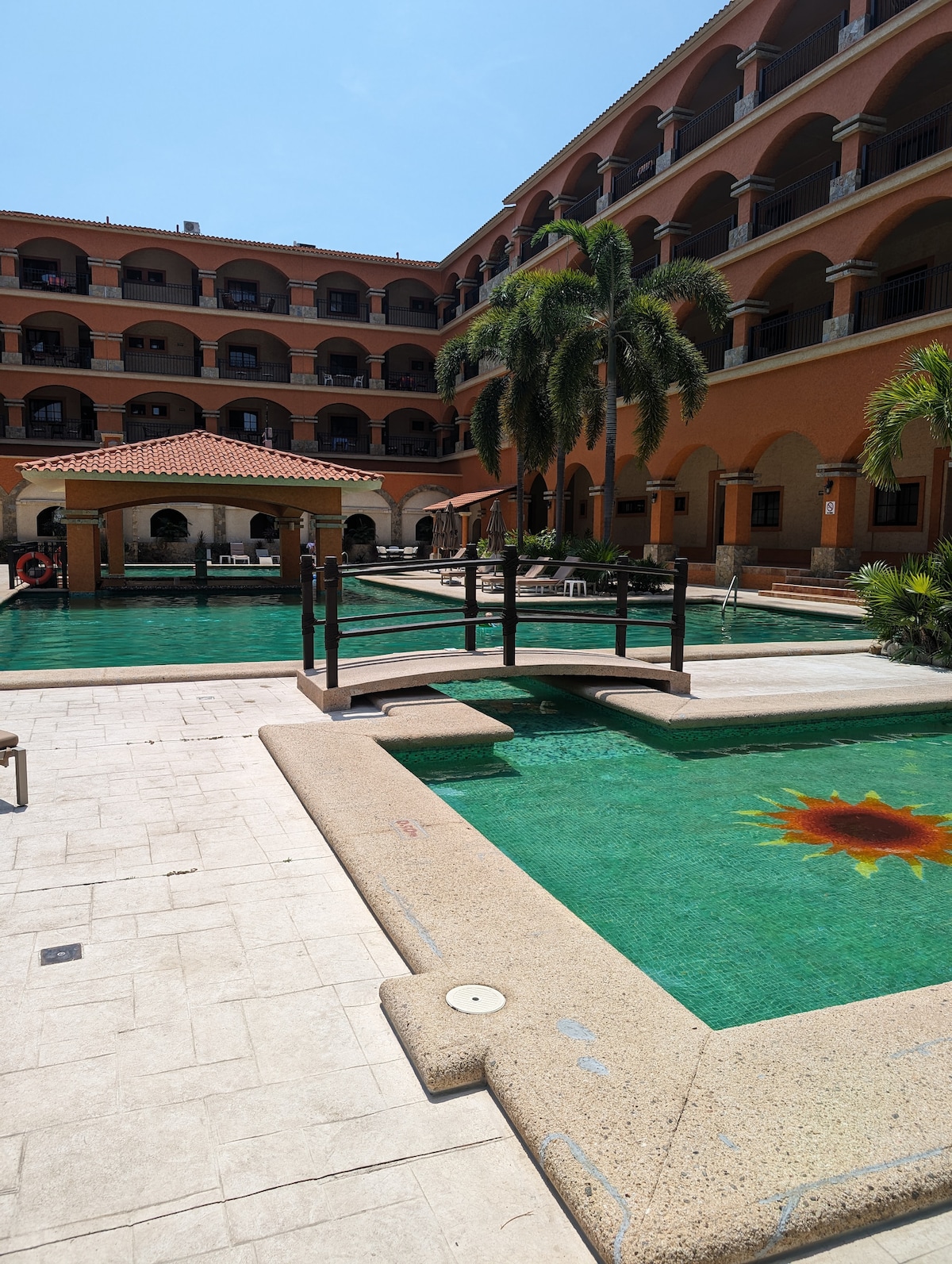 The pool area features multiple swimming sections surrounded by well-maintained landscaping. Sun loungers are placed along the edges, and a wooden bridge connects different sections. The building's architecture creates a harmonious backdrop, with balconies overlooking the water.