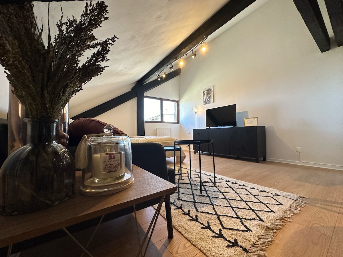 A bright living area is featured with sloped ceilings and wooden beams. A comfortable sofa is positioned next to a sleek television stand. A small coffee table sits on a textured rug, with a decorative vase and a candle adding a touch of elegance to the space.