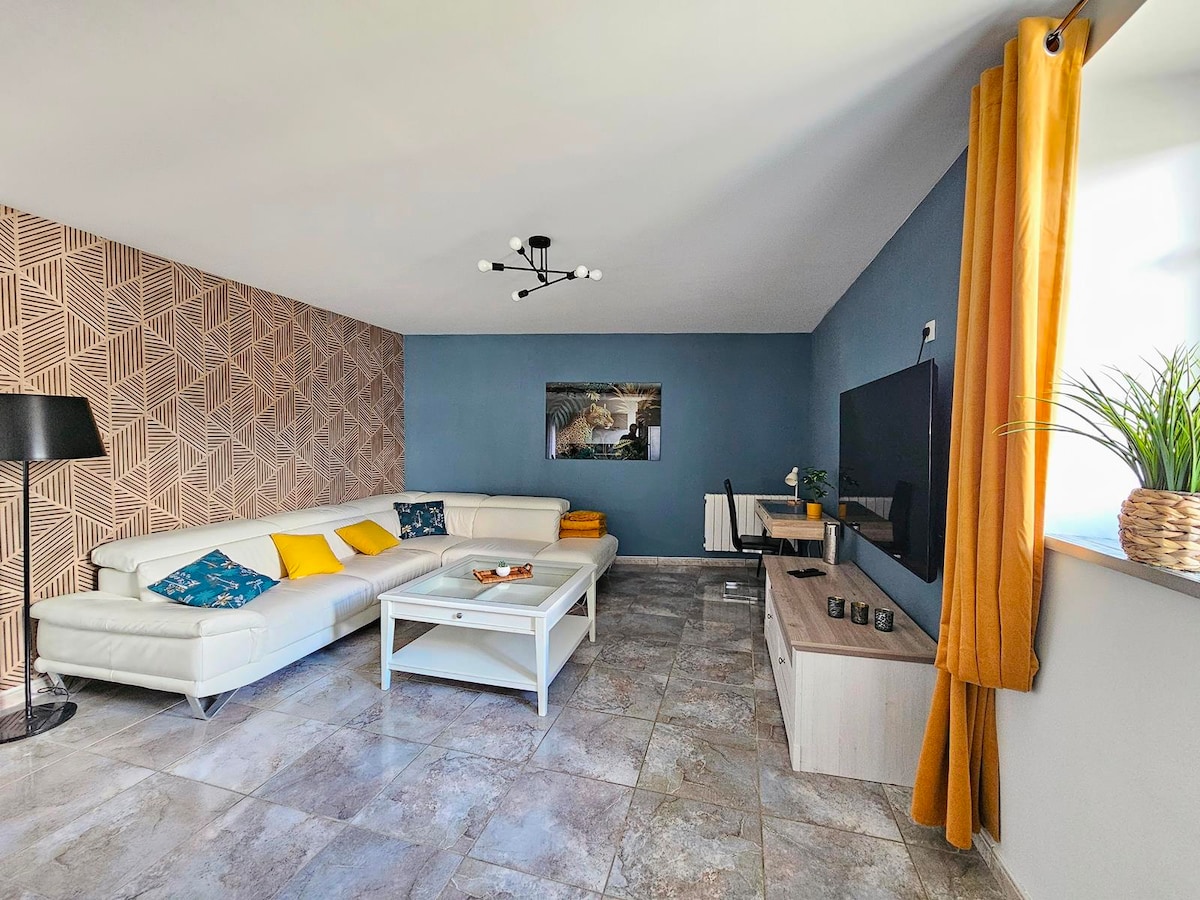 A spacious living area features a white L-shaped sofa adorned with decorative pillows in shades of blue. A light wood coffee table sits in front, while a flat-screen TV is mounted on a wall opposite a large window dressed with yellow curtains. A decorative plant adds a touch of greenery.