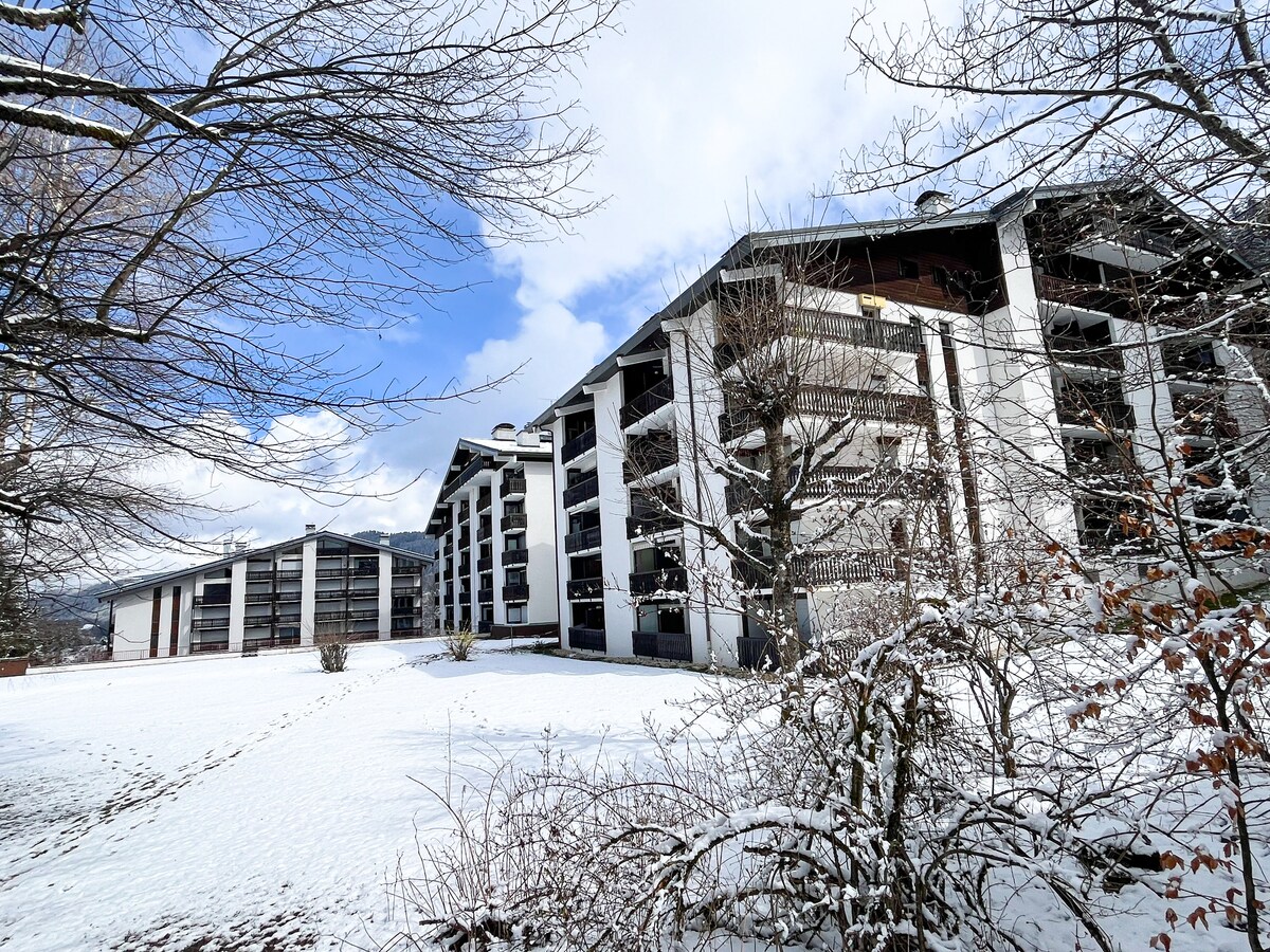 The image depicts two residential buildings in a serene, snow-covered landscape. Clear blue skies contrast with the white snow. The architecture features multiple balconies and large windows, surrounded by soft white snow and a few leafless trees, enhancing the peaceful setting.