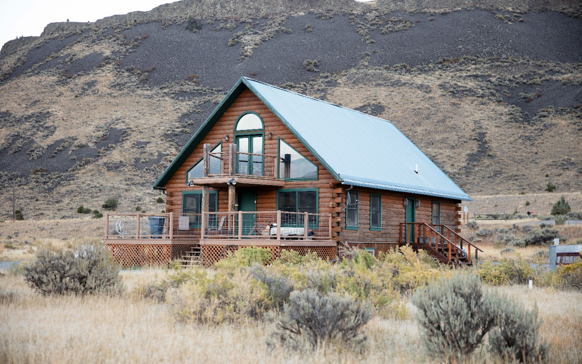 A rustic log cabin with a teal metal roof is surrounded by natural grassland and hills. The spacious front deck features railings and steps leading to the ground. Large windows reflect the surrounding landscape, inviting natural light into the home.