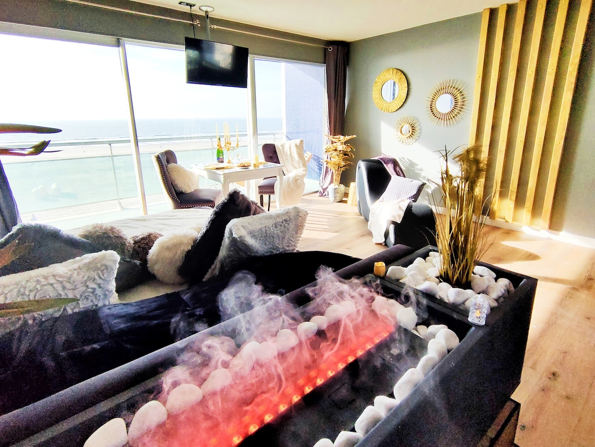 A modern living space is depicted, featuring a sleek electric fireplace with glowing embers surrounded by smooth stones. Large windows offer views of the ocean, while stylish seating and a dining table are visible. Decorative mirrors and a touch of greenery enhance the ambiance.