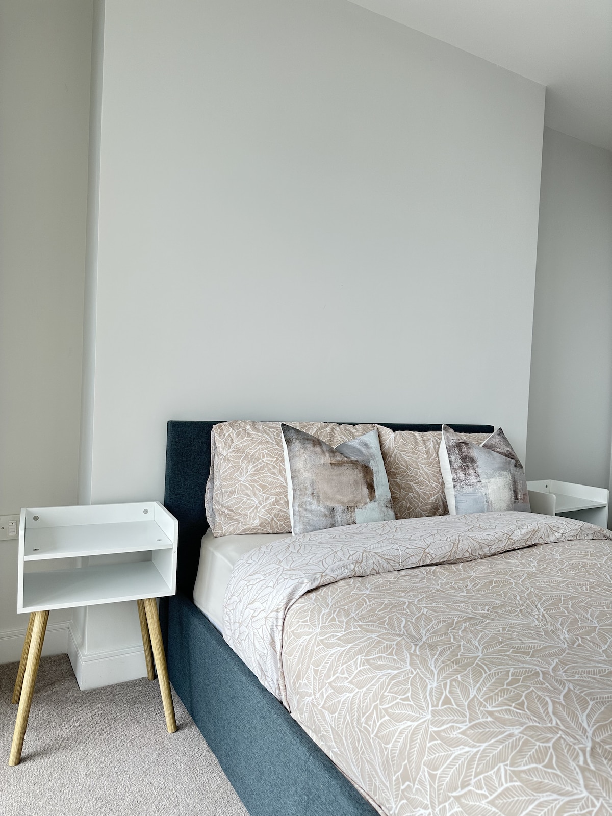 A welcoming bedroom features a spacious bed with a light-toned patterned duvet and decorative pillows. Two minimalist nightstands with sleek designs flank the bed, enhancing the modern aesthetic. Soft, neutral tones contribute to a calming atmosphere.