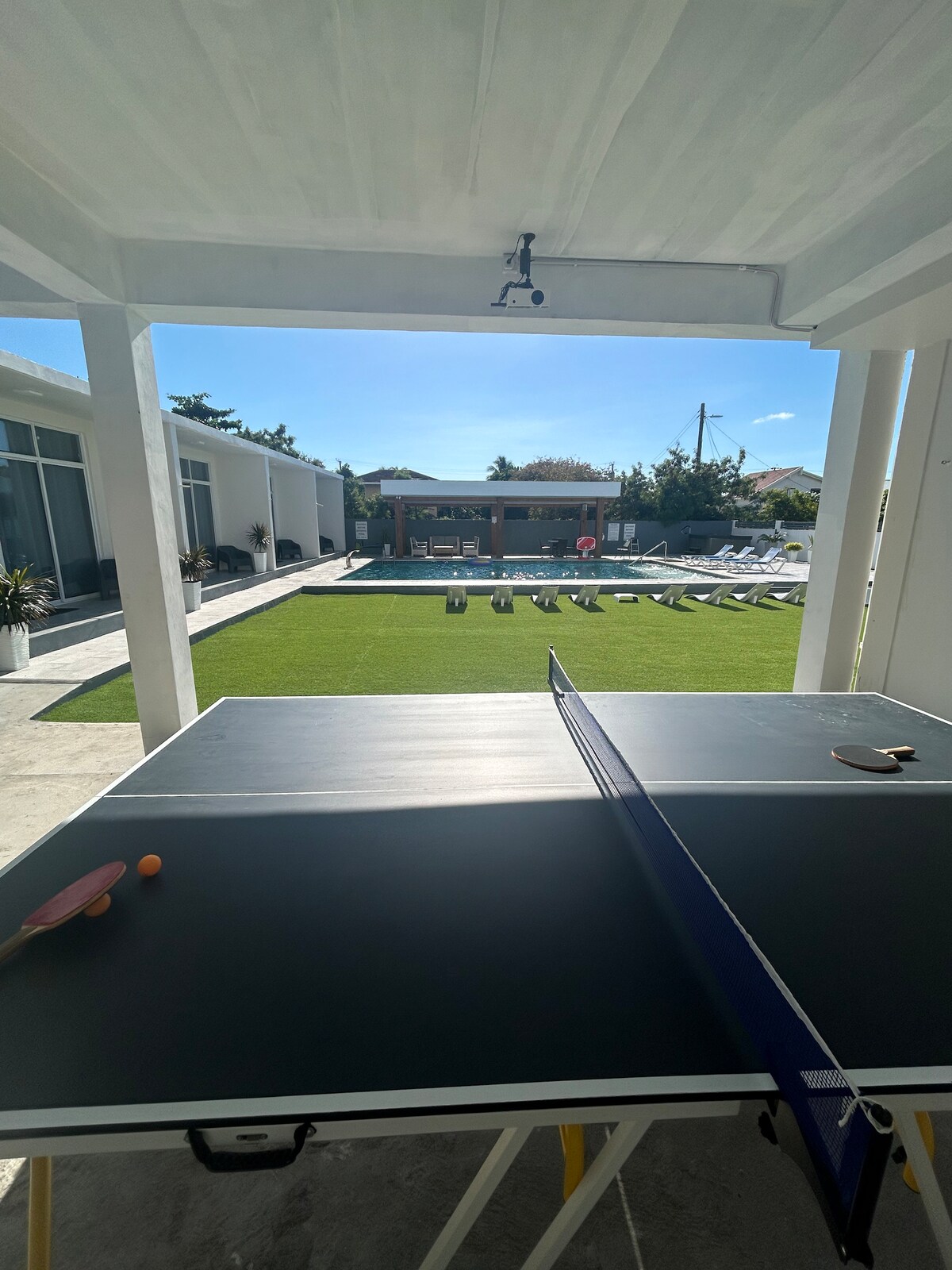 A ping pong table is positioned in the foreground, overlooking a lush green lawn and a swimming pool. Sun loungers are arranged neatly around the pool area, while bright sunlight illuminates the outdoor space, creating a vibrant atmosphere.