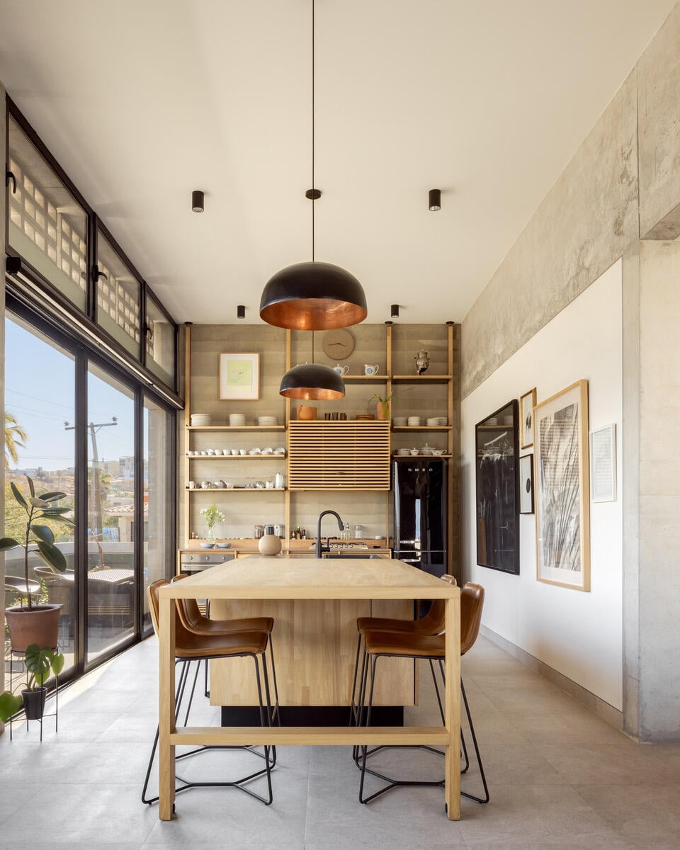 The kitchen features a large, light wood island topped with a smooth surface, surrounded by subtle metal stools. Two pendant lights hang above, illuminating the space. Open shelving displays neatly arranged dishware, and floor-to-ceiling windows provide ample natural light and views of the outside.