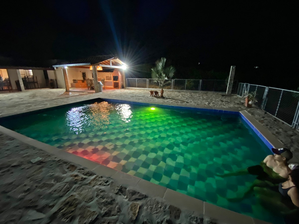A private pool is illuminated at night, with colored lighting reflecting off the water's surface. A covered outdoor area is visible in the background, along with a well-lit kitchen space. A stone patio surrounds the pool, creating a relaxed environment.
