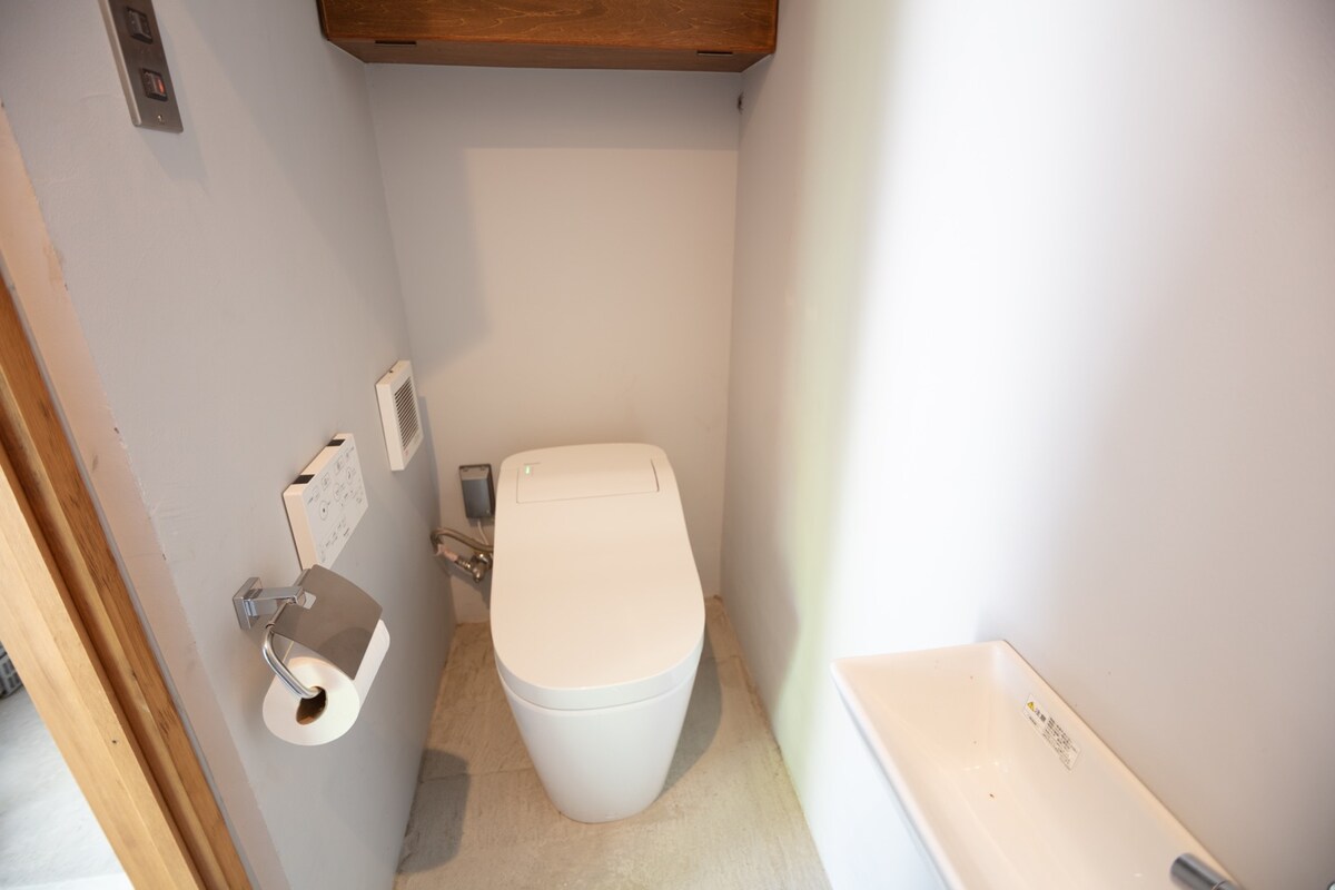 A modern restroom is showcased, featuring a sleek toilet design with a side-mounted flush panel. A small washbasin is situated next to the toilet, accompanied by a roll of toilet paper on a wall-mounted holder. The soft, neutral walls create a clean, uncluttered appearance.