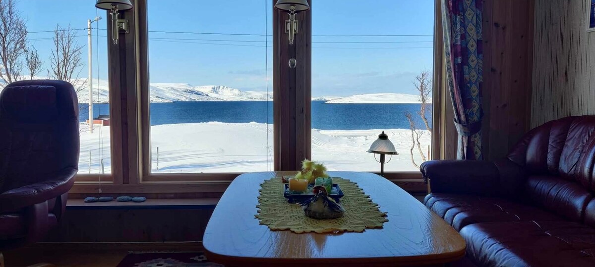A bright living area features large windows overlooking a snow-covered landscape and a serene body of water. Comfortable seating options are arranged around a wooden table, which displays a decorative cloth with various items. Soft natural light enhances the inviting atmosphere.