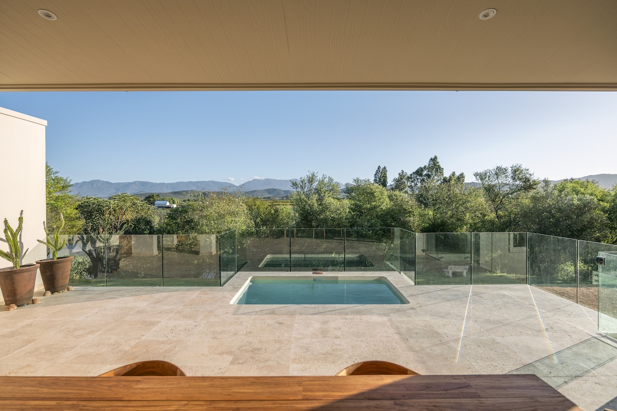 A spacious outdoor area is visible, featuring a swimming pool surrounded by glass fencing. The view extends to lush greenery and distant mountains, while a wooden table is positioned to overlook the tranquil scene.
