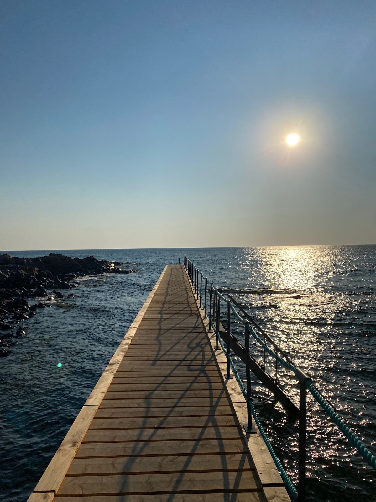 A wooden pier extends into the shimmering water, leading toward the horizon where the sun is positioned overhead. The surface of the water glistens, reflecting the sunlight while rocky formations border the edges of the scene.
