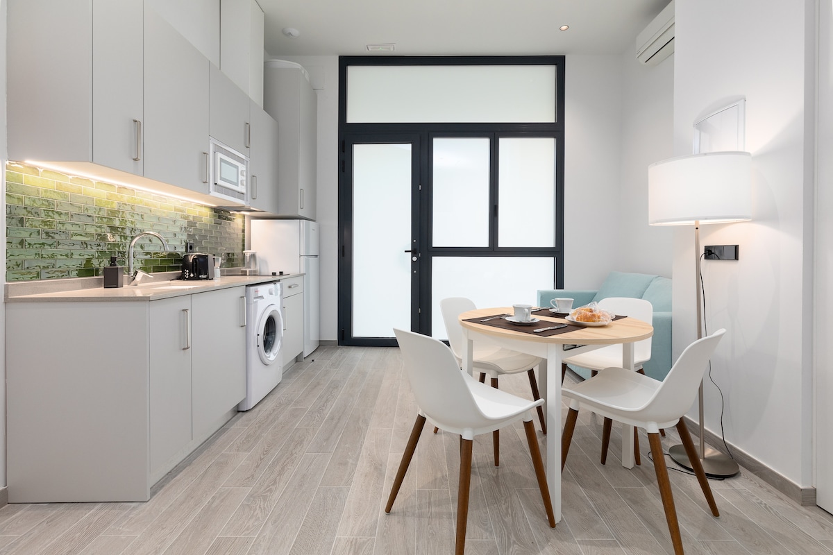 A modern kitchen is featured with sleek cabinetry and a microwave, set against a green-tiled backsplash. A compact dining table is positioned near a window, surrounded by four white chairs. A light-colored sofa is present in the corner, enhancing the open layout.