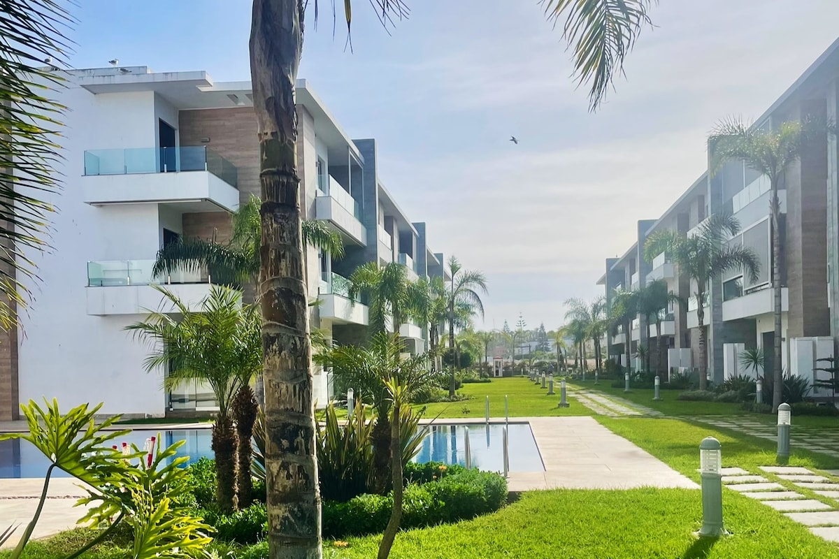 A peaceful outdoor area is framed by well-maintained gardens featuring lush greenery and palm trees. Paths lead toward low-rise buildings with modern architecture, while a rectangular pool reflects the surrounding landscape, complemented by a manicured lawn.