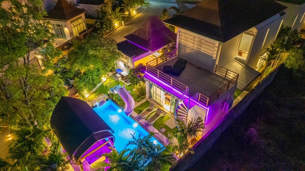 An aerial view captures a private pool villa, surrounded by lush greenery and illuminated by soft lighting. A serene pool glimmers at the center, flanked by lounging chairs and outdoor spaces. The modern architecture features a balcony, enhancing the overall tranquil environment.