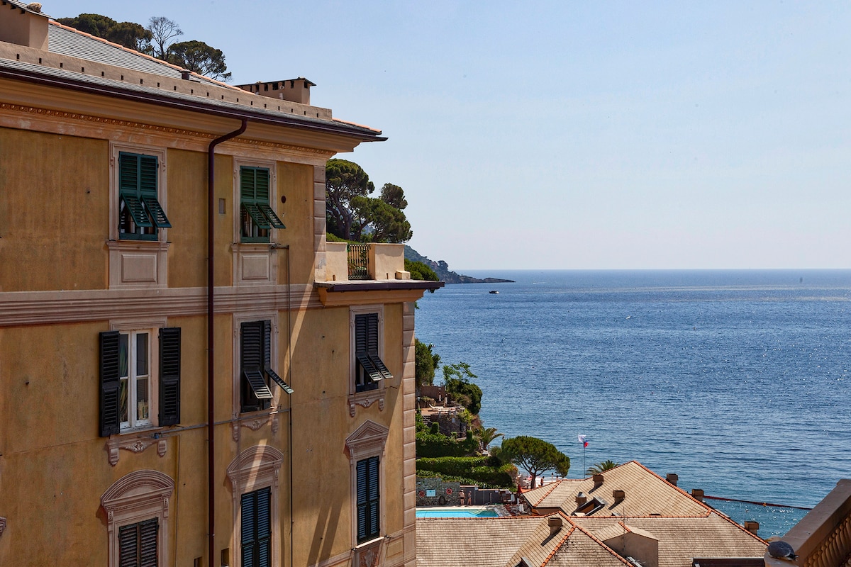 An expansive view of the Mediterranean coastline is presented, featuring calm blue waters and a sandy beach. The scene includes historic buildings with green shutters that reflect the area’s architectural style, framed by lush greenery and the distant shoreline.