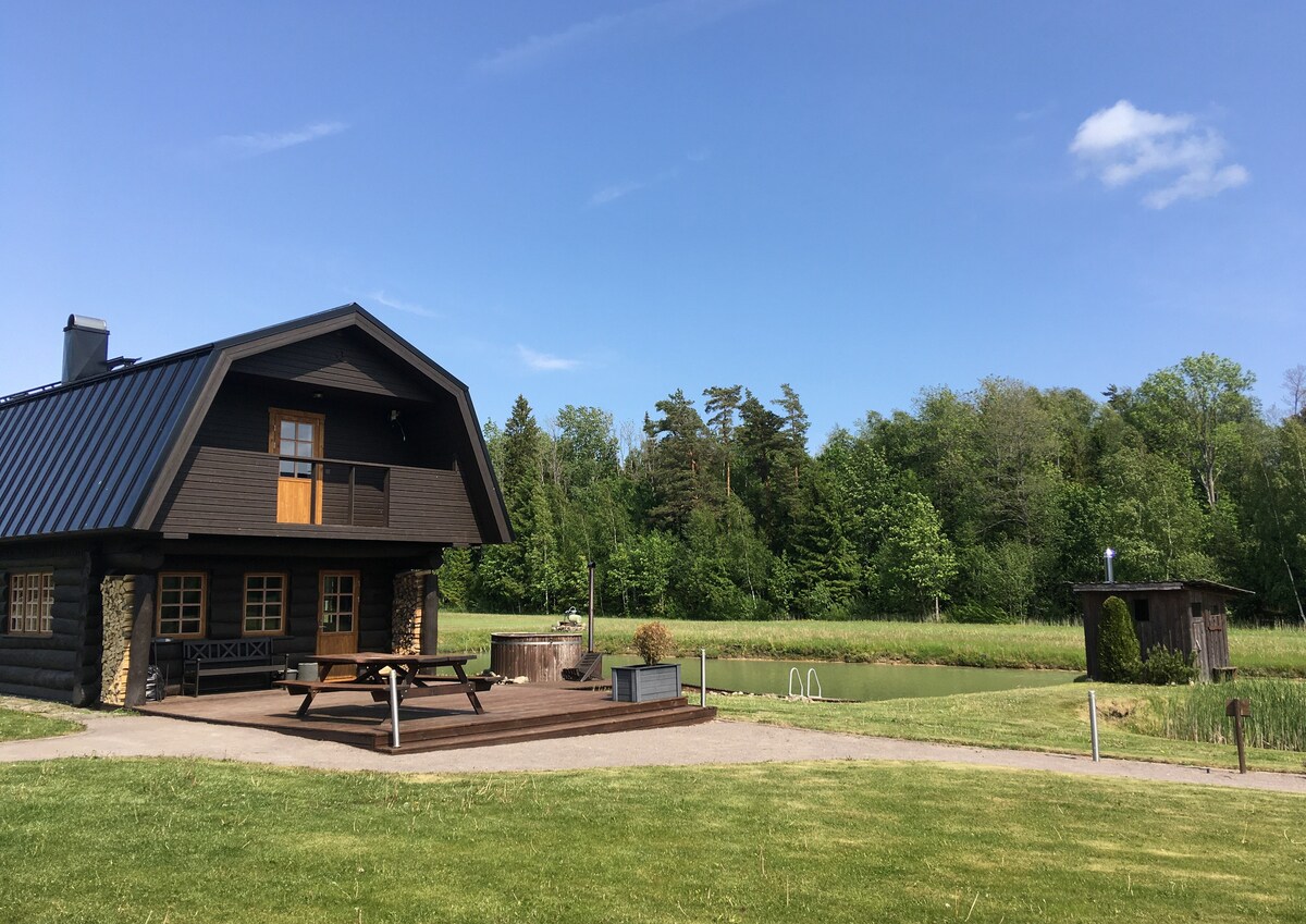 A dark wooden house with a sloped roof is set near a serene pond. A spacious deck features a picnic table for outdoor dining. Surrounding greenery and trees provide a natural backdrop, with a small outbuilding visible nearby.