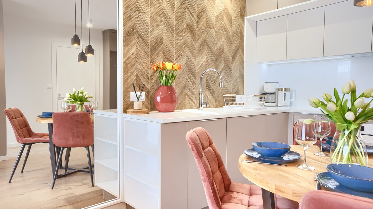 A modern kitchen and dining area feature sleek cabinetry and a herringbone patterned accent wall. A round wooden table is set with blue plates and glassware, surrounded by plush pink chairs. Fresh flowers add a touch of color to the space.
