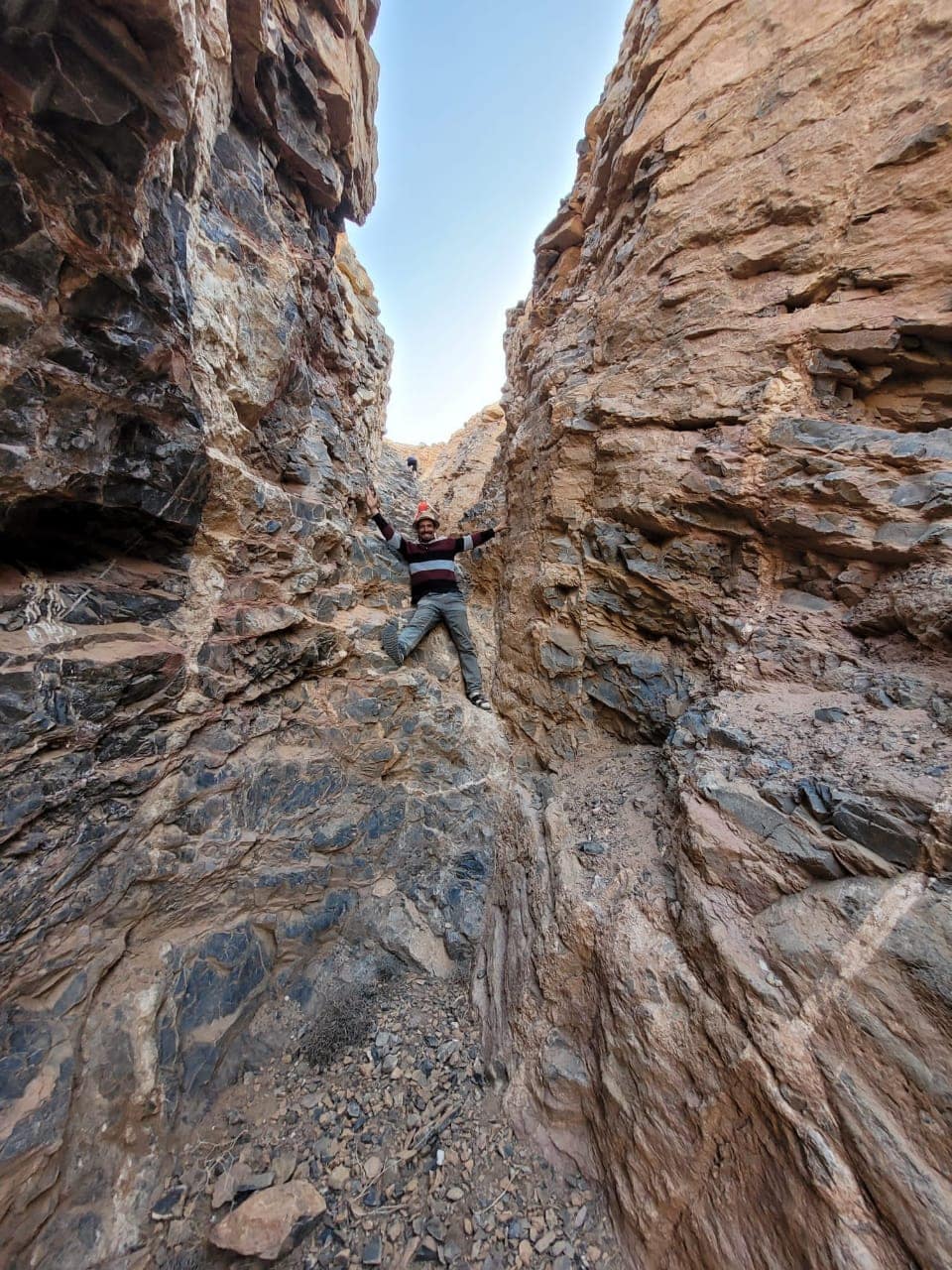 A narrow canyon is depicted, with steep rocky walls rising on either side. A person is positioned within the canyon, appearing to climb against the rugged terrain. Sunlight illuminates the orange and brown stone, highlighting the texture and shadows throughout the landscape.