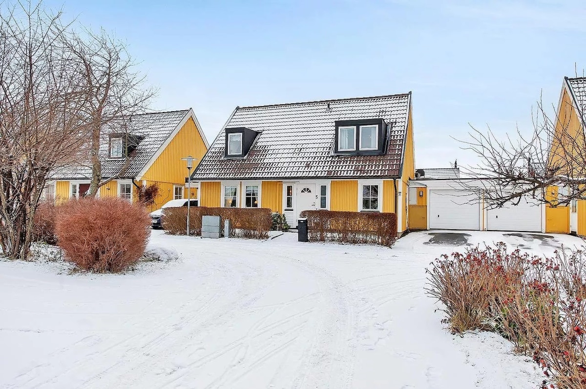 A charming yellow house with a sloped roof stands prominently on a snowy landscape. Surrounding greenery is visible, including shrubs and trees. A driveway leads to the house, with a garage on the right. Snow covers the ground, providing a tranquil winter scene.