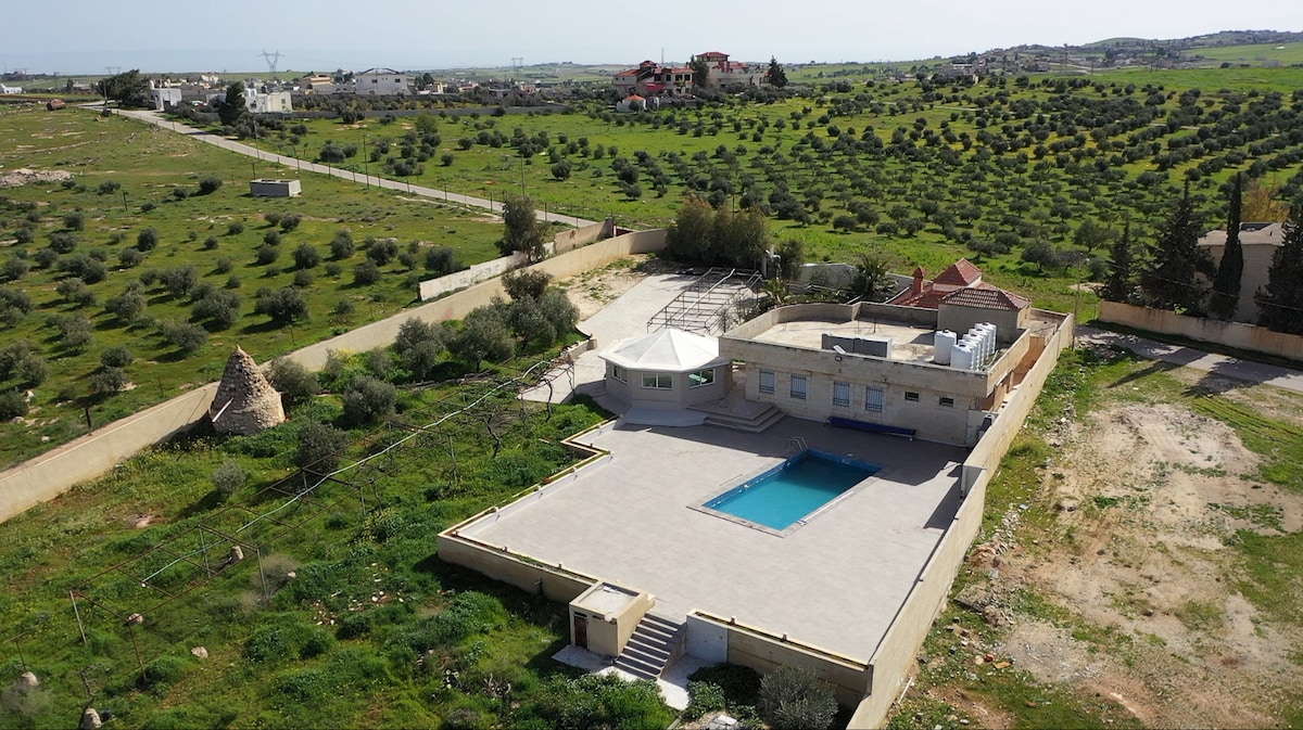 An aerial view captures a spacious property surrounded by lush greenery and rolling hills. A rectangular pool is centered on the expansive patio, which is equipped with outdoor seating and shaded areas. The home's neutral tones complement the peaceful landscape.